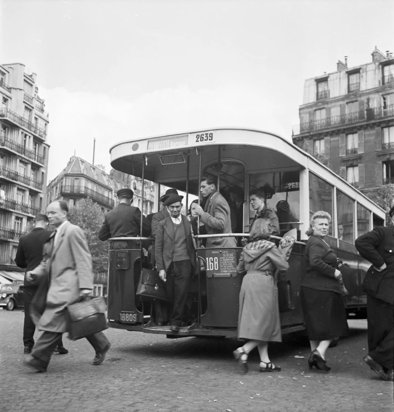 Un autobus Renault TN à plate-forme de la ligne 168, avec passagers, dans une rue parisienne bordée d’immeubles haussmanniens